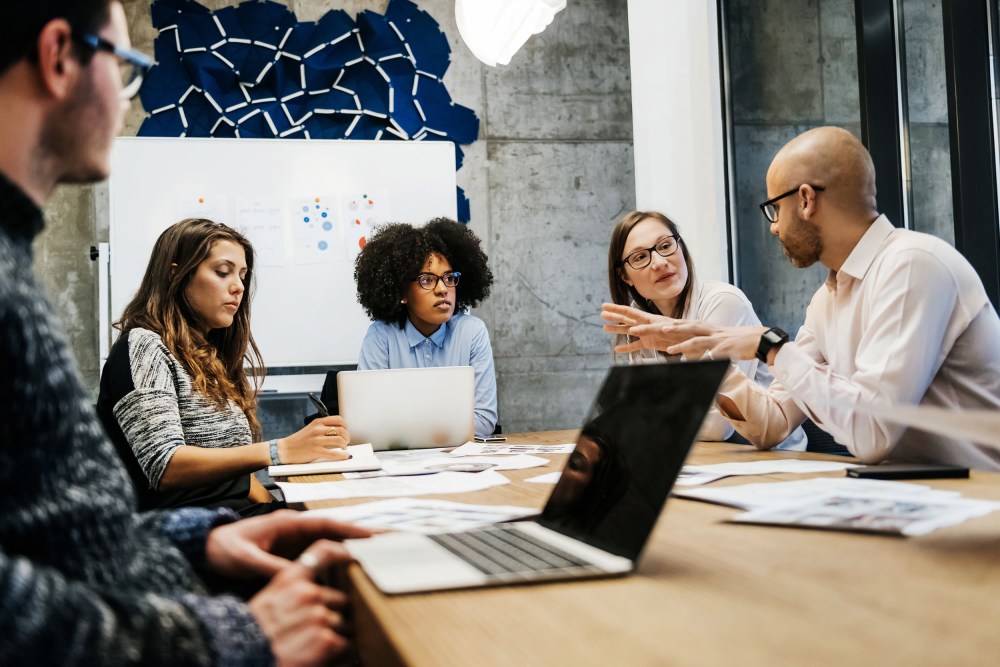 Image: A group of three young women and two men of different ethnicities hold a business meeting in a modern day office.