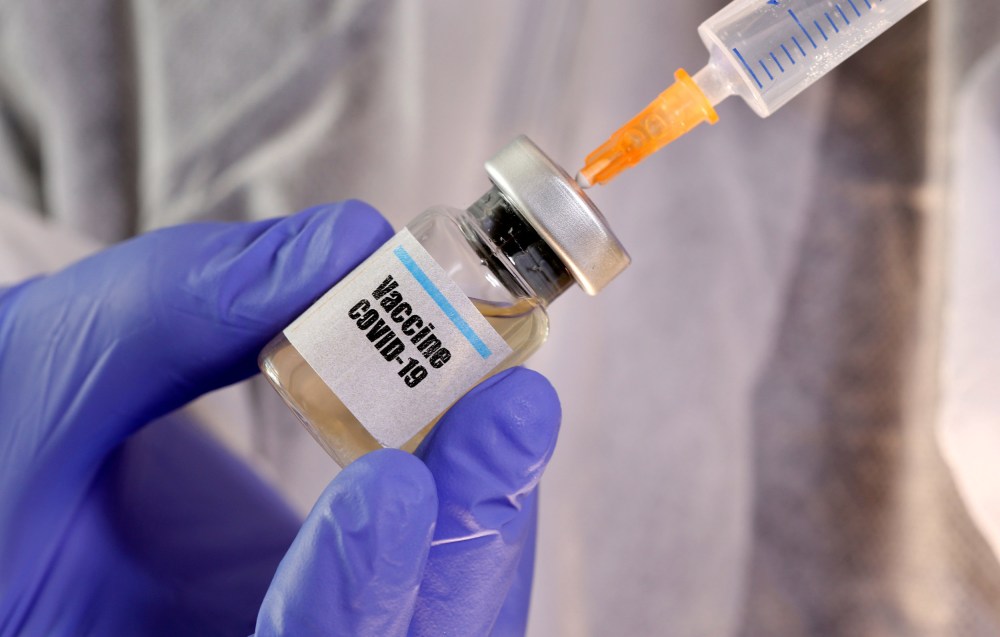 Image: A woman holds a small bottle labeled with a "Vaccine COVID-19" sticker and a medical syringe.