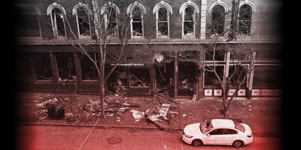 Image with a red overlay of debris remains on the sidewalk in front of buildings damaged in an explosion. A car is parked on the right.