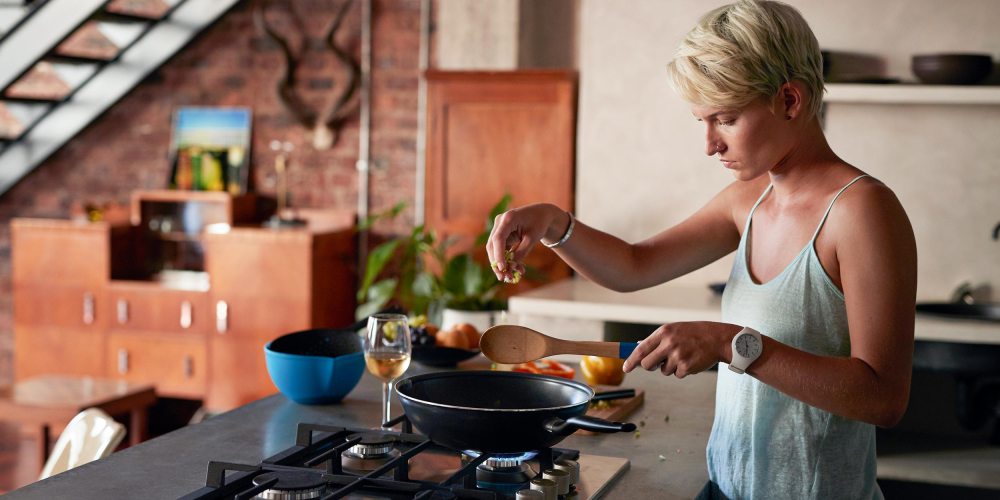 Woman standing in her kitchen cooking with a pan. Shop the best Mother's Day gifts for foodie moms and moms who love cooking. The best Mother's Day kitchen gifts include cutting boards, pans and more.