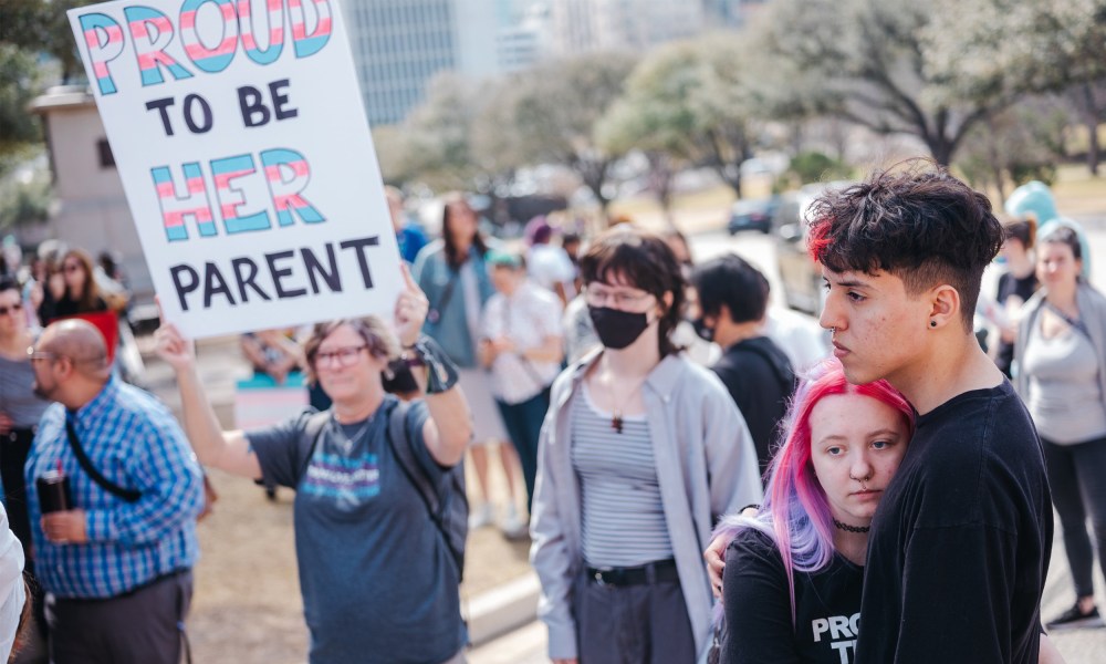 Image: A demonstrator holding a sign that reads,"Proud to be be her parent". The words, "Proud" and "her" are in the transgender pride flag colors.