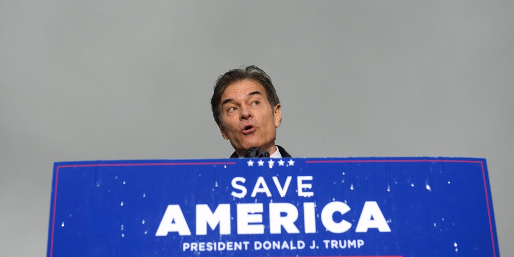 Image: Dr. Mehmet Oz standing behind a podium with a sign that reads,"Save America, President Donald j. Trump".