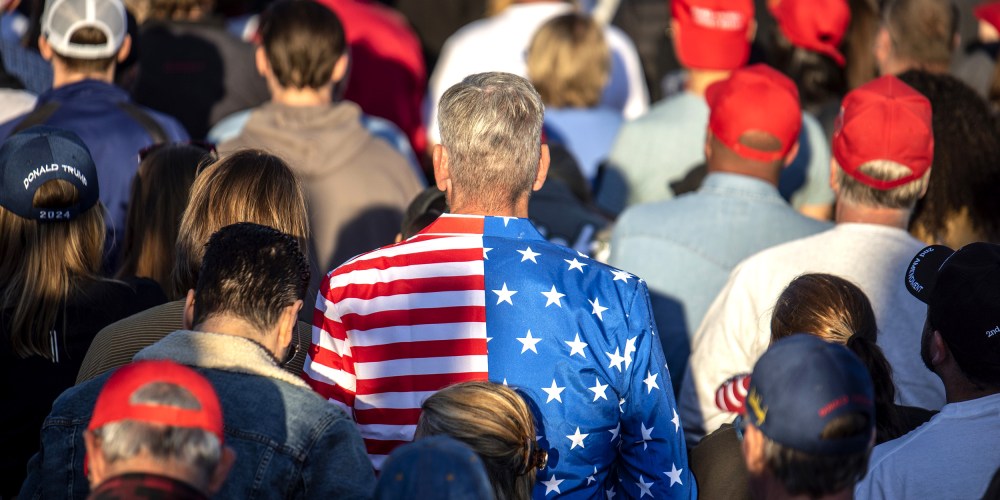 Image: Man in the centre wearing a blazer with American flag colors amidst attendees at a rally.