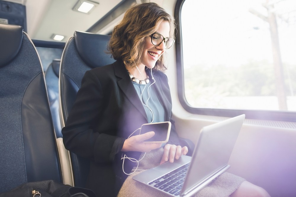 Mid adult woman on train, using smartphone and laptop