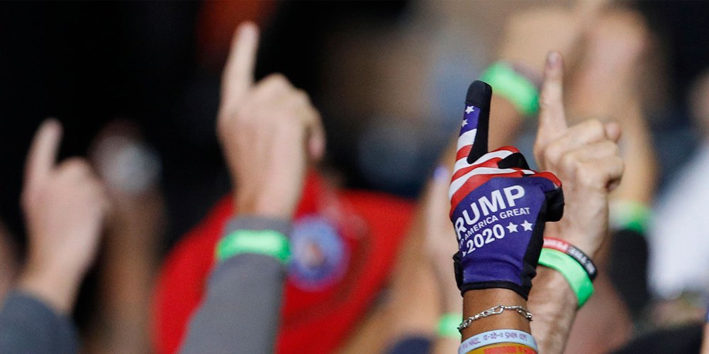 Image:People at a rally holding their index fingers up. One of the hands is shown wearing a blue and red glove that reads,"Trump 2020".