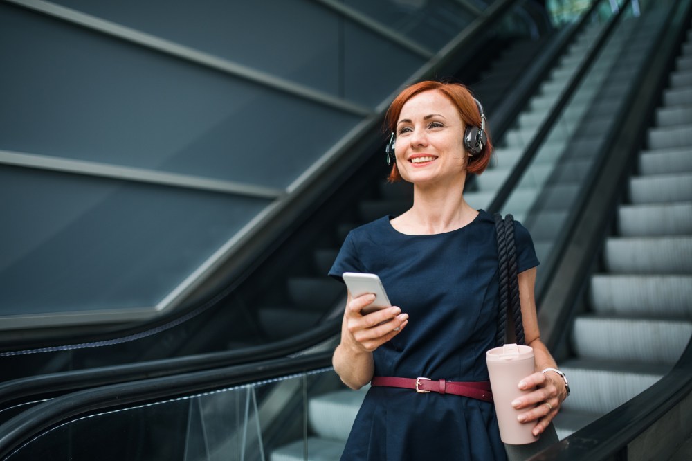 Young businesswoman with coffee and smartphone indoors on escalator in city, listening to music.