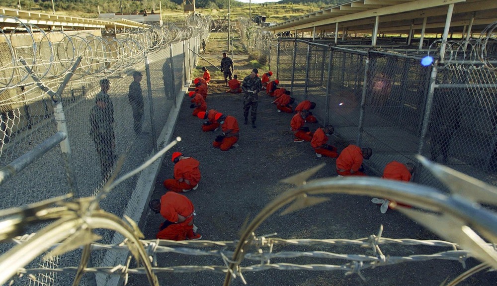 Detainees in orange jumpsuits sit in a holding area under the watchful eyes of military police during in-processing to the temporary detention facility at Camp X-Ray of Naval Base Guantanamo Bay in this January 11, 2002 file photograph.