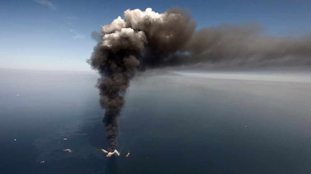 In this Wednesday, April 21, 2010 file photo, oil can be seen in the Gulf of Mexico, more than 50 miles southeast of Venice on Louisiana's tip, as a large plume of smoke rises from fires on BP's Deepwater Horizon offshore oil rig.