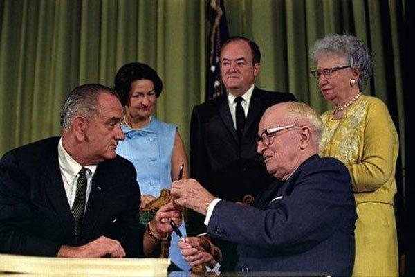 LBJ prepares to sign Medicare into law. Former President Harry Truman hands him the pen, as VP Hubert Humphrey, Lady Bird Johnson and Bess Truman look on.