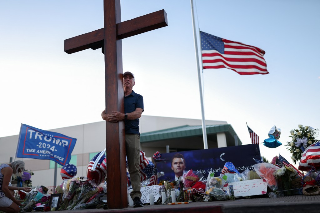 A man holds a giant cross by a memorial for Charlie Kirk.