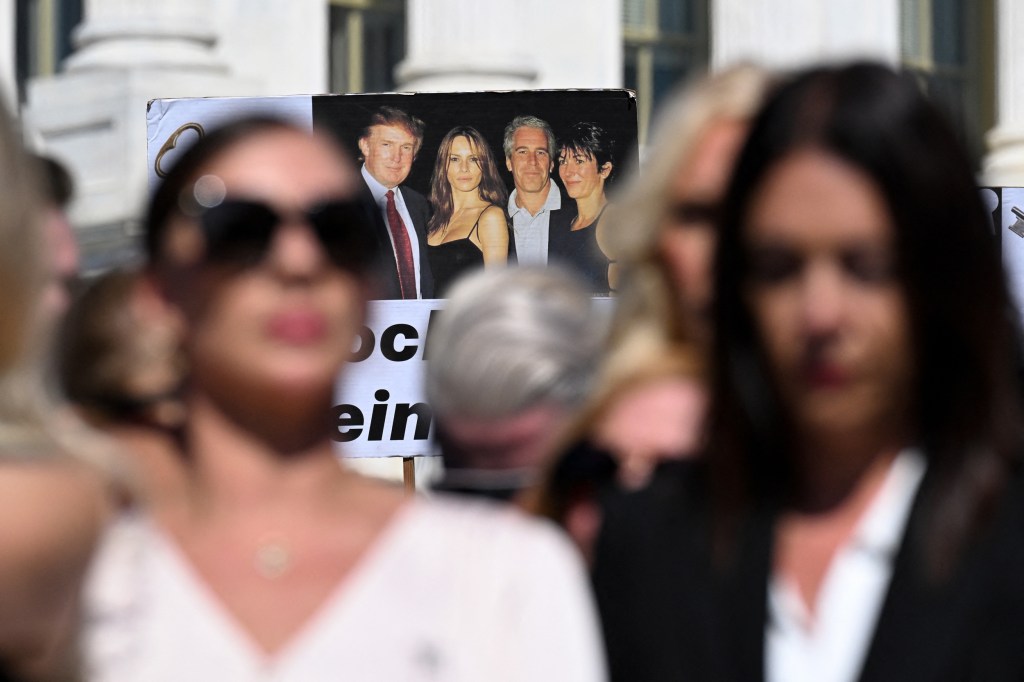 A poster featuring a photo of Donald Trump, Melania Knauss, Jeffrey Epstein and Ghislaine Maxwell in focus behind people attending a press conference and rally in support of the victims of sex offender.