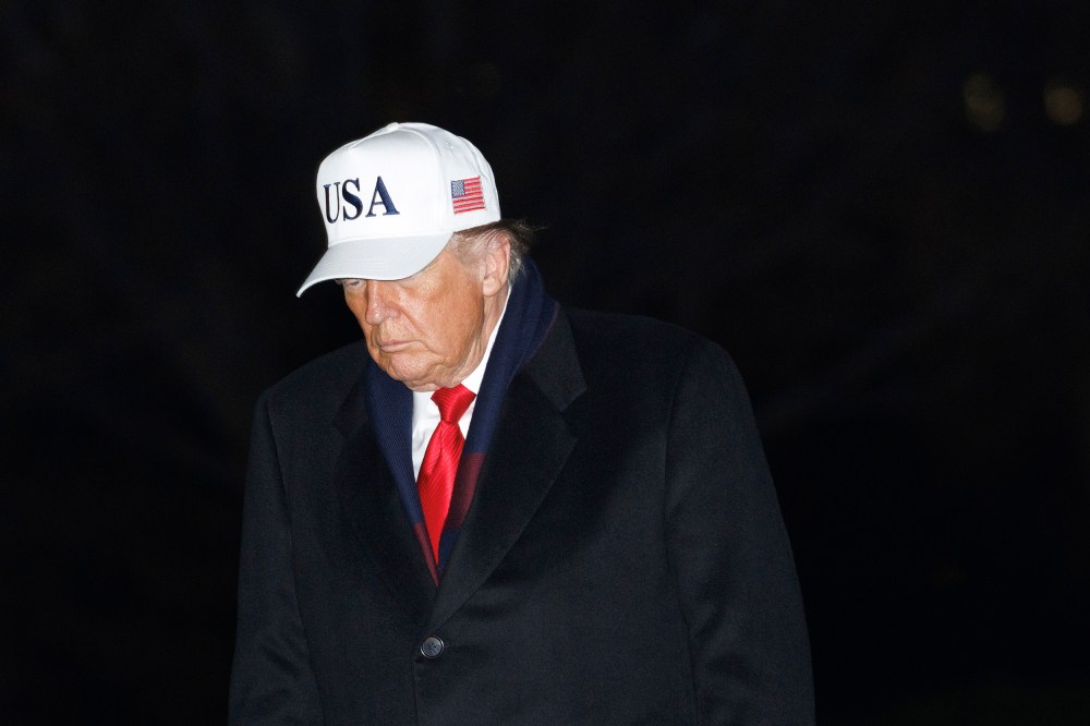 Donald Trump walks at the White House at night, wearing a white USA hat.