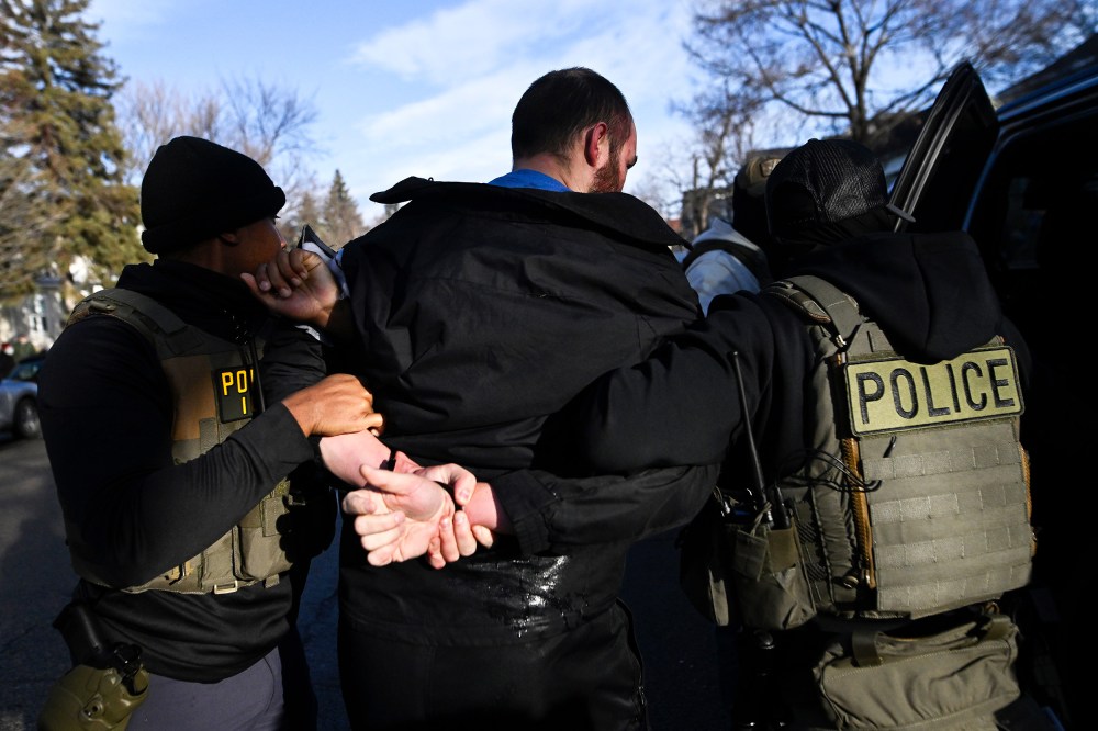 A person with their hands restrained behind their back, surrounded by people in vests labeled "police."