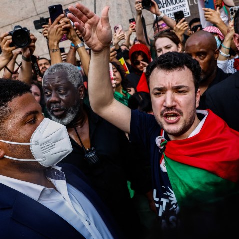 Mahmoud Khalil raises his hand while protesting with a big crowd. A Palestinian flag is draped over his shoulder.