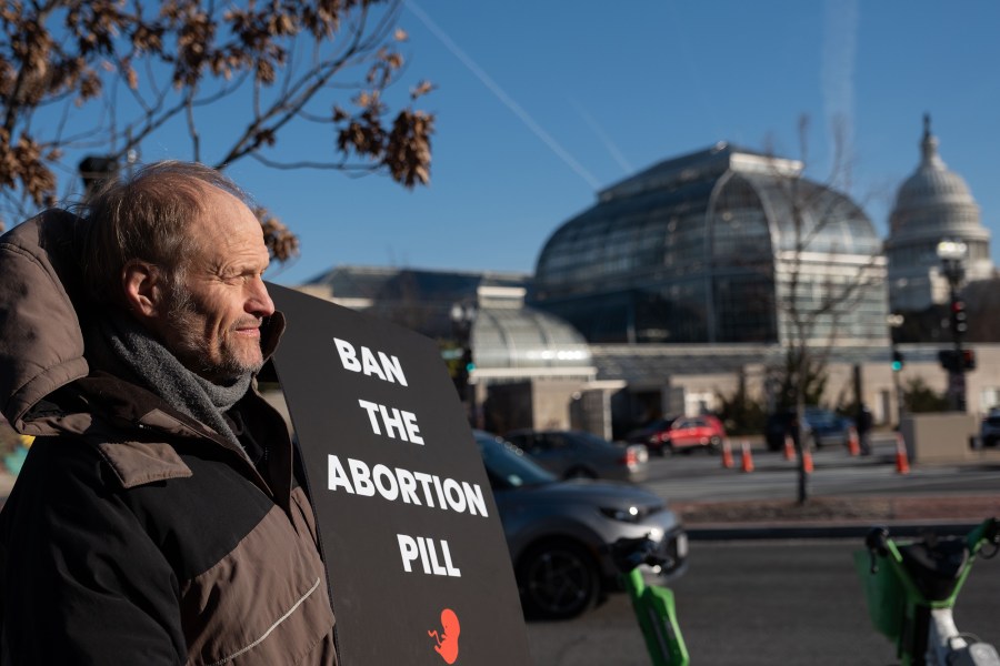 John Hinshaw stands with a sign that reads "Ban the abortion bill" on the left of the frame. The Capitol can be seen in the background. 