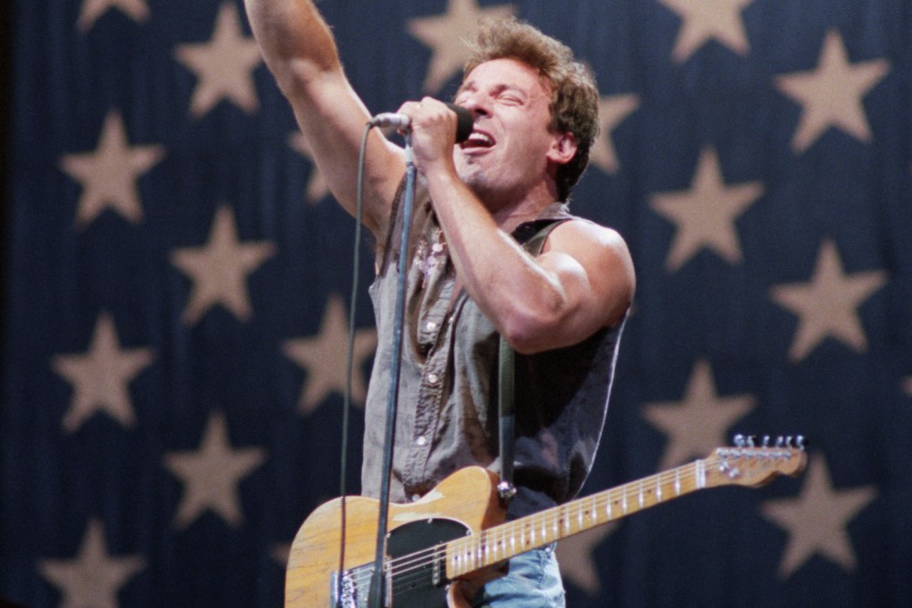 Bruce Springsteen with his guitar on stage, in front of a giant American flag.