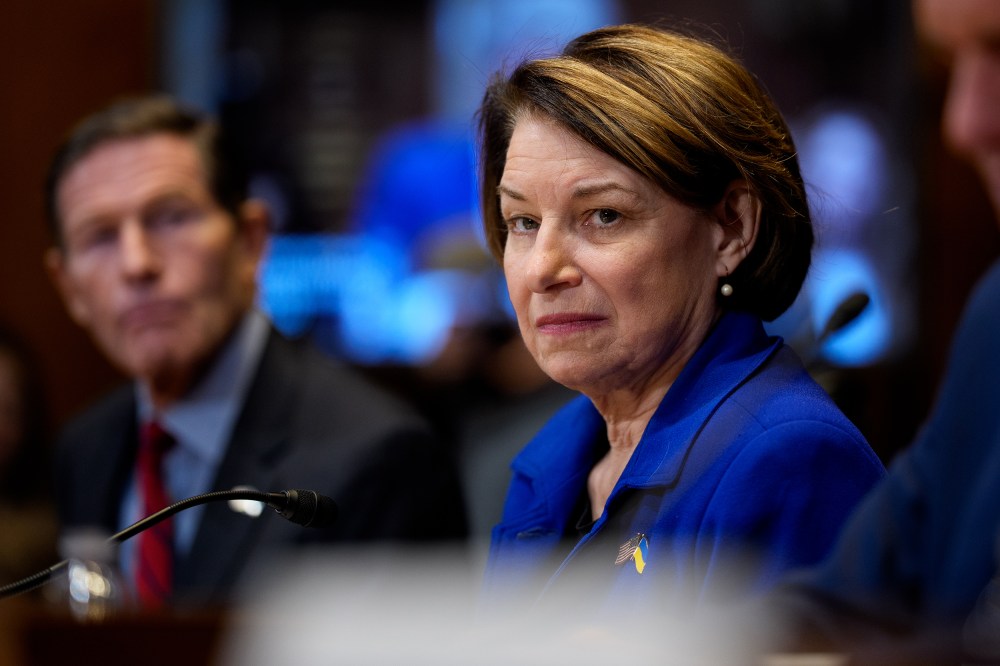 Sen. Amy Klobuchar sitting between other senators while they testify- wearing a bright blue jacket.