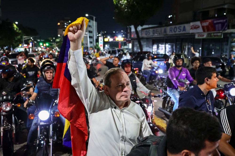 Juan Pablo Guanipa, center, stands in the street, holding a Venezuelan flag up with one hand. He is surrounded by people on motorcycles.