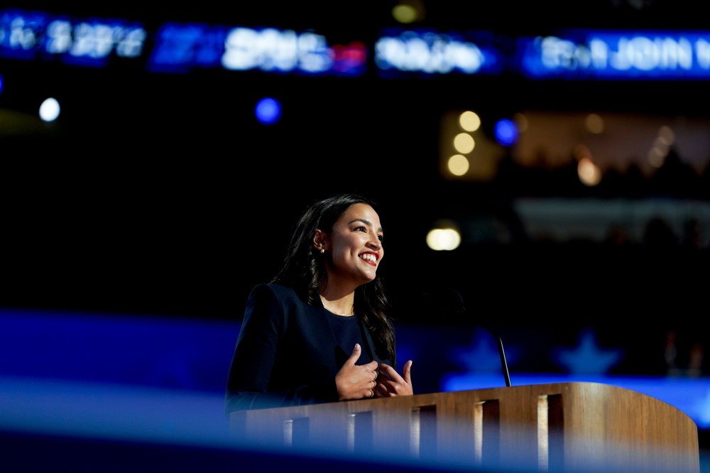 Alexandria Ocasio-Cortez smiles while standing behind a podium.