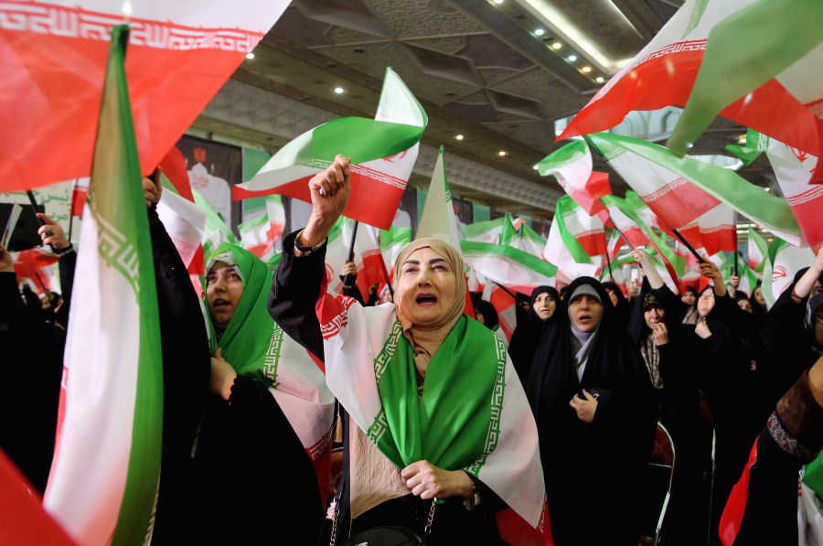 One women raises her right hand with an Iranian flag thrown over her shoulders - surrounded by many other women wearing hair covers and carrying the country's flag.