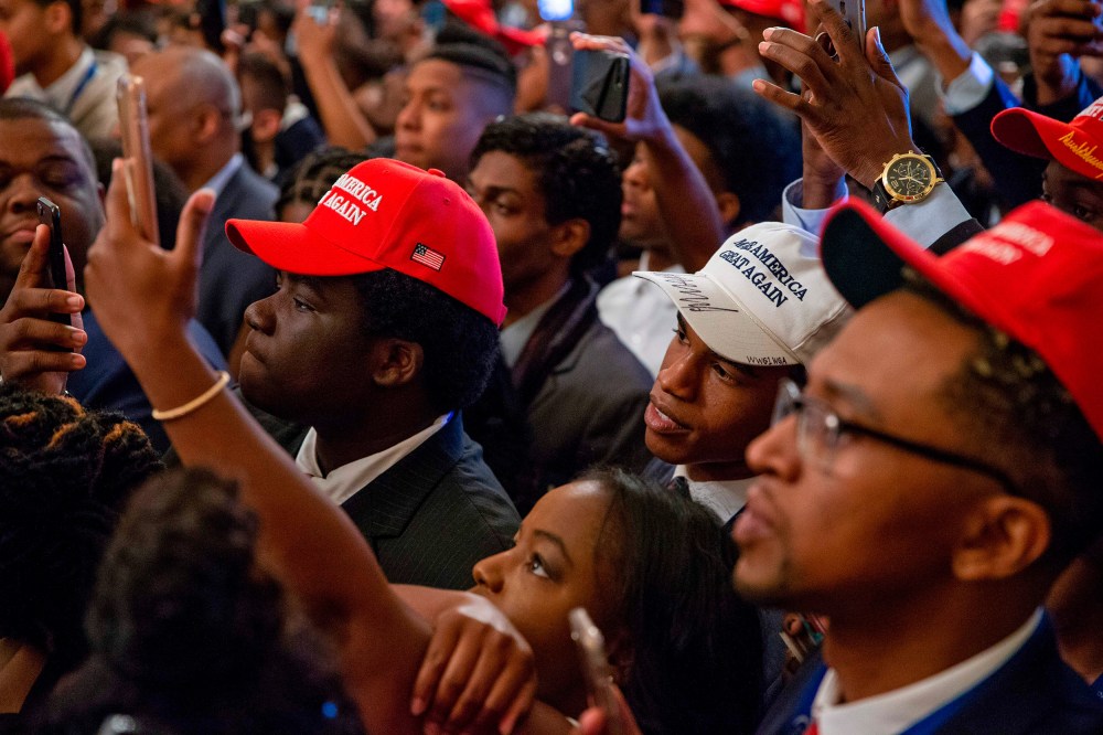 Audience members, most of them Black, wear MAGA hats and hold up their phones to take photos.