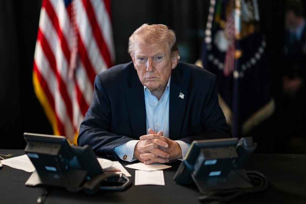 Donald Trump sits at a table with two phones in front of him and U.S. flags behind him.