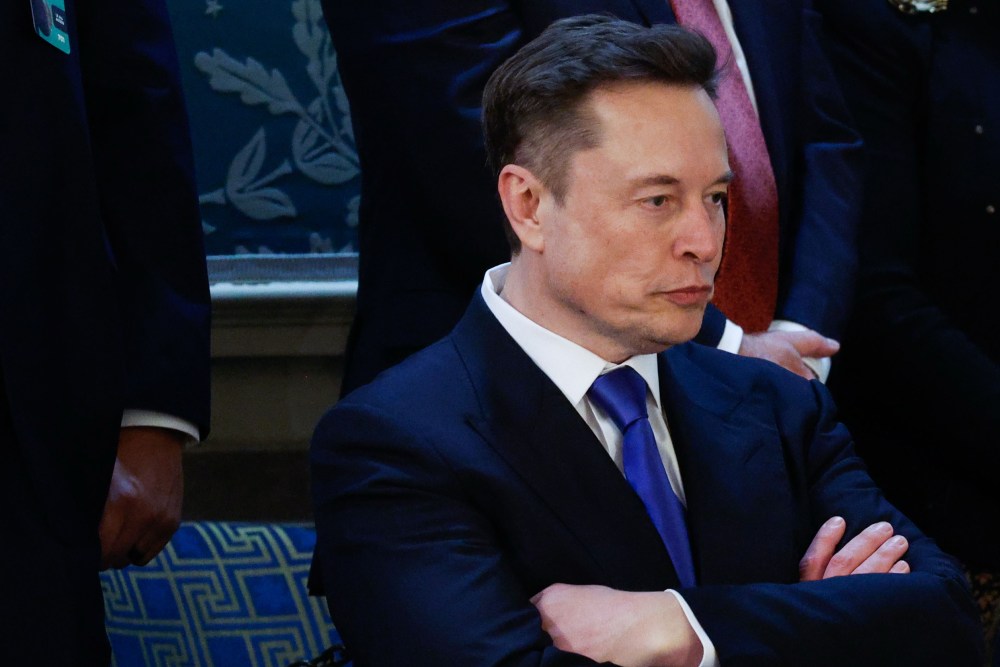 Elon Musk looks down and has his arm crossed on his chest.