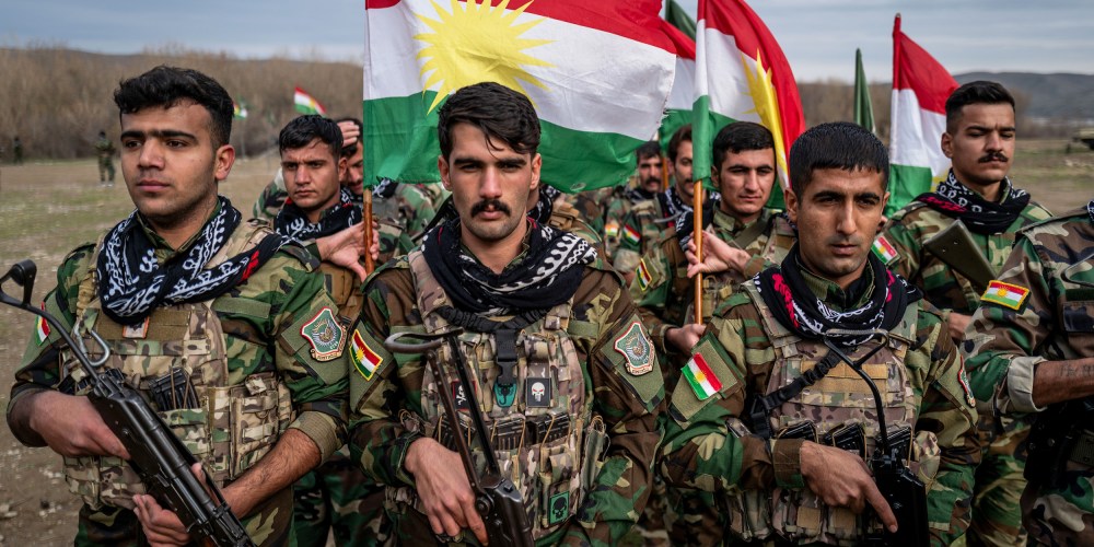 People in military uniforms holding guns and Kurdish flags stand in formation.