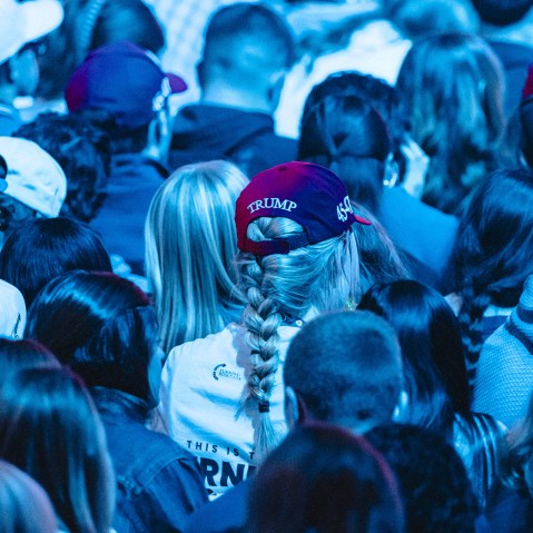 Backs of people's heads sitting in an audience. Multiple people wear Trump hats. The entire photo is tinted blue by the lights.