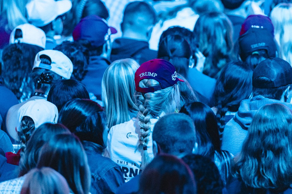 Backs of people's heads sitting in an audience. Multiple people wear Trump hats. The entire photo is tinted blue by the lights.