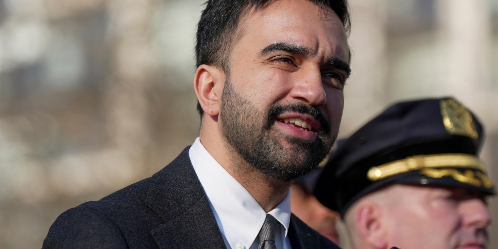 In this tight shot, Zohran Mamdani is seen speaking in front of a mic while a law enforcement officer stand behind to the right.