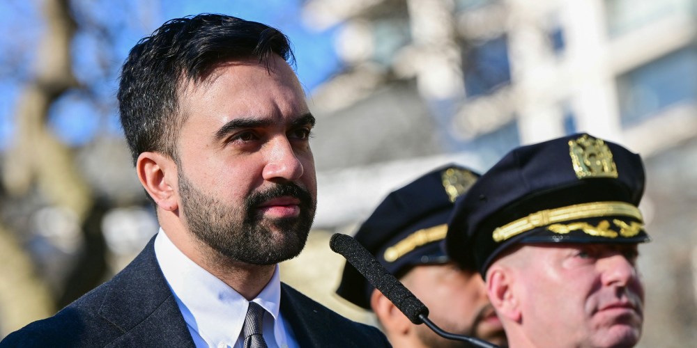 Zohran Mamdani stands in front of a microphone. Police officers are seen behind him.
