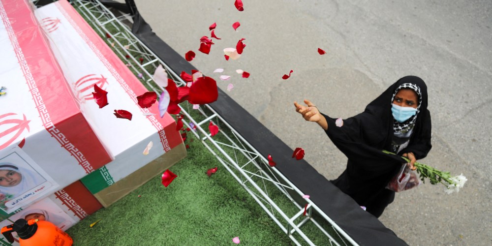 A woman is seen wearing a black hijab on the right side of the frame as she throws flower petals on a coffin.