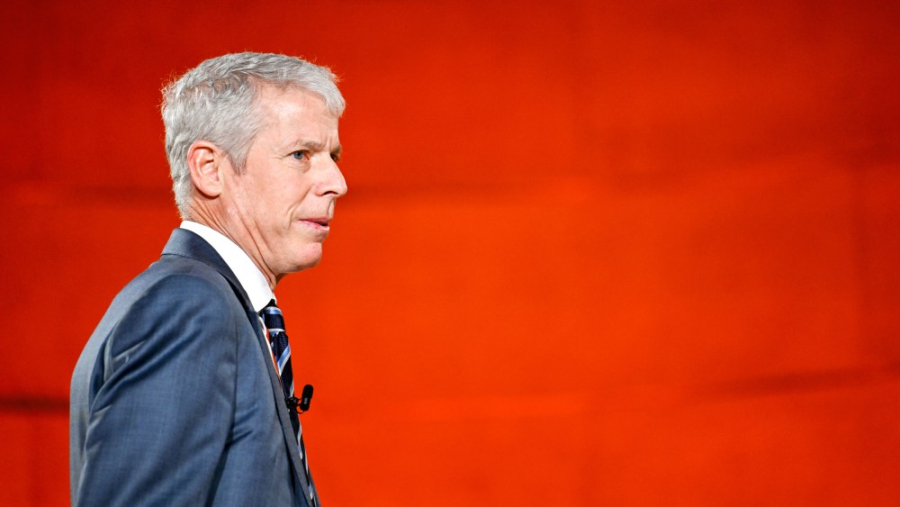 Chris Wrights walks towards right side of the frame in front of a red background.