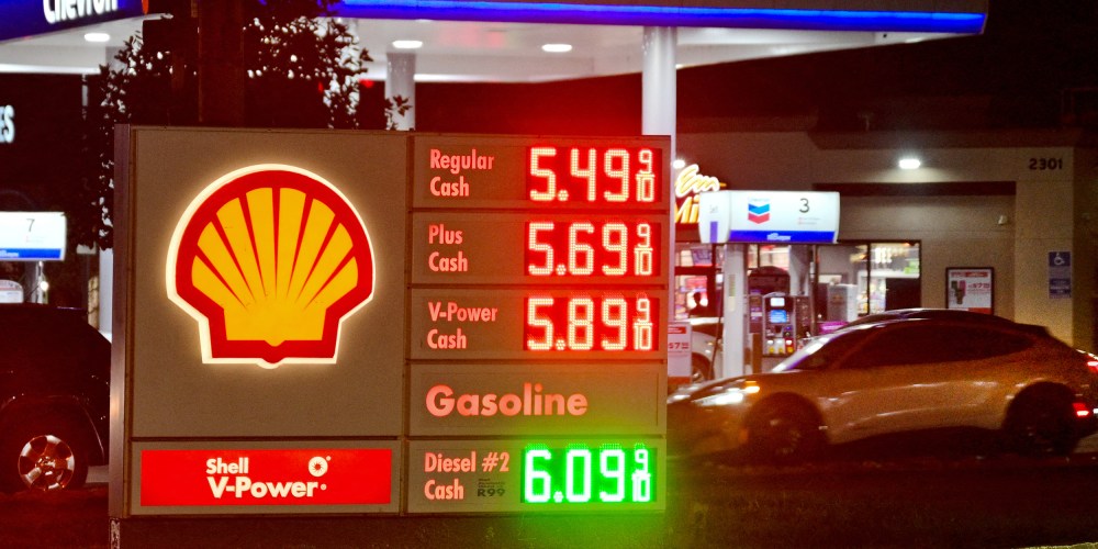 A gas station sign on the ground shows fuel prices while a car pulls into to get gas.